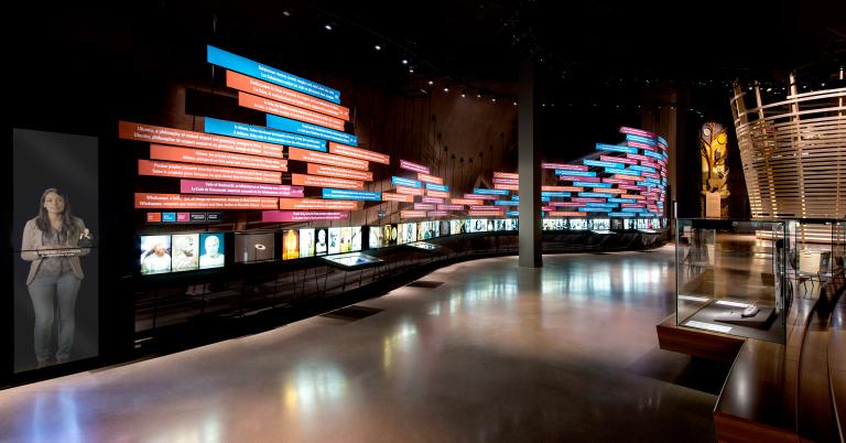 Vue d’une galerie muséale. On voit du texte sur des rectangles colorés sur un grand mur d’exposition qui s’étend sur toute la longueur de la galerie. Sous cette chronologie d’événements se trouvent des photos rétro-éclairées en rangée. Sur le côté droit de la photo, on voit un grand élément d’exposition montrant une scène de nature et une structure circulaire en bois. Visibilité masquée.