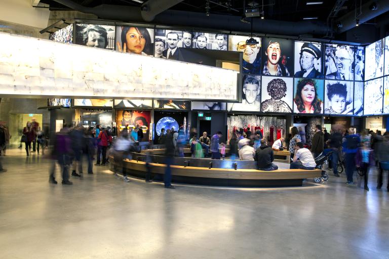 Une grande foule dans une galerie de musée. Il y a un banc courbe au centre de la salle. Dans l’espace, on voit une série de grandes images placées au-dessus d’alcôves qui contiennent chacune une exposition différente.