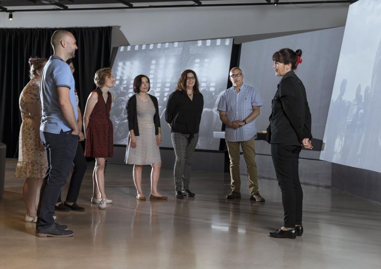 Un petit groupe de gens debout en demi-cercle devant de grands écrans inclinés. Visibilité masquée.