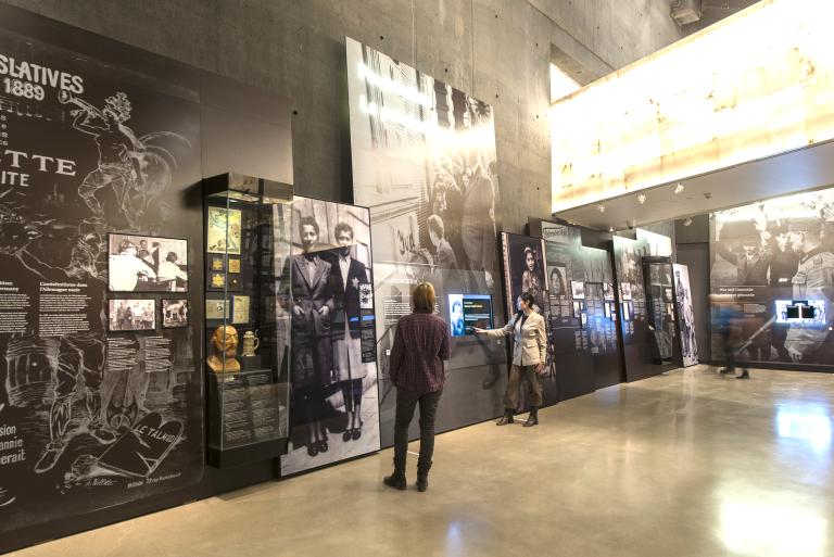 Deux personnes explorent une galerie de musée contenant des images, des objets, du texte et des écrans interactifs.