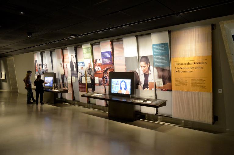 Deux personnes interagissent avec l’un de deux écrans vidéo dans une galerie de musée. Il y a des images de gens et des blocs de texte en couleur sur le mur.
