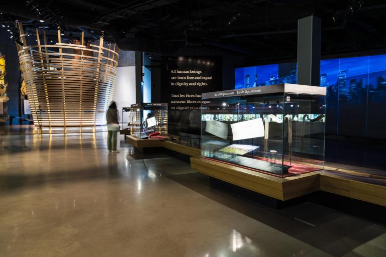 Une galerie de musée contenant une structure circulaire formée de lattes horizontales de bois, des vitrines contenant des objets et des projections sur le mur. Visibilité masquée.