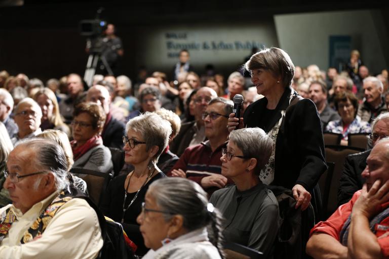 Une salle remplie de personnes assises. Une femme est debout et tient un microphone portable. Visibilité masquée.