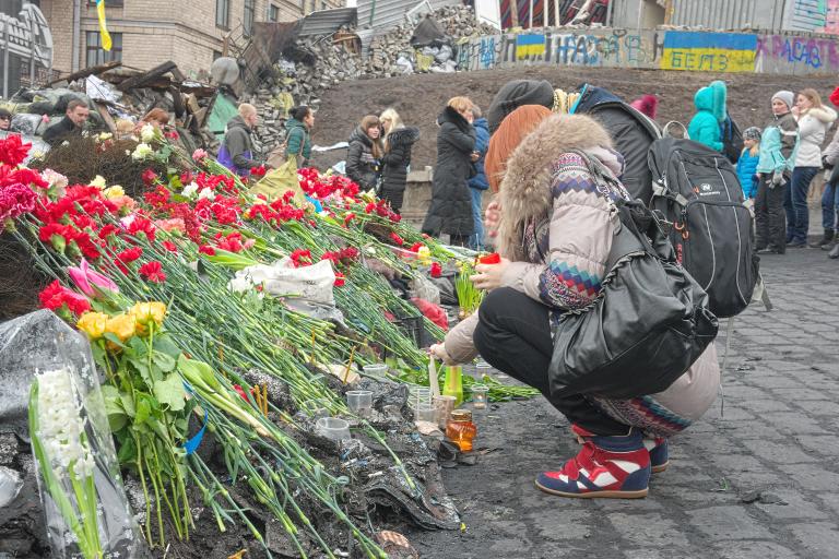 Deux personnes déposant des fleurs rouges à un mémorial. En arrière-plan, une douzaine d’autres personnes se tiennent devant des débris, comme des roches, des feuilles de métal et des barrières de ciment peintes avec des drapeaux ukrainiens bleu et jaune et des lettres. Visibilité masquée.