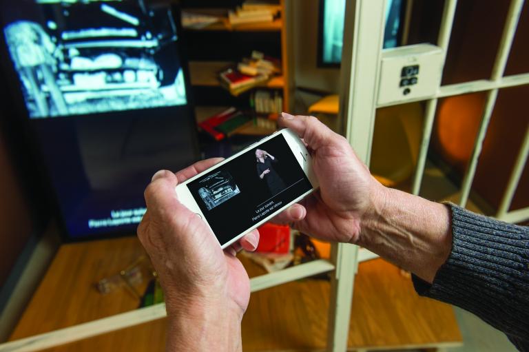 Deux mains tiennent un téléphone intelligent devant une exposition muséale. Sur l’écran du téléphone, on aperçoit une femme qui fait de l’interprétation en langage gestuel. On voit aussi des sous-titres en français.