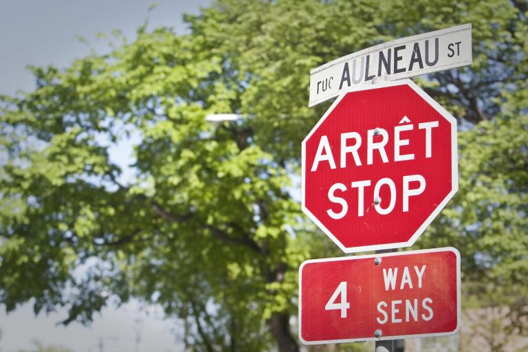 Un panneau d’arrêt et un panneau de signalisation de rue, en français et en anglais, se trouvent devant des arbres. Visibilité masquée.