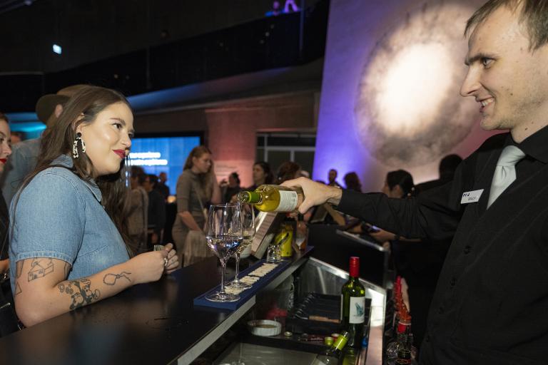 A woman waits for a drink at a bar in a museum space, while a bartender pours glasses of wine. Visibilité masquée.