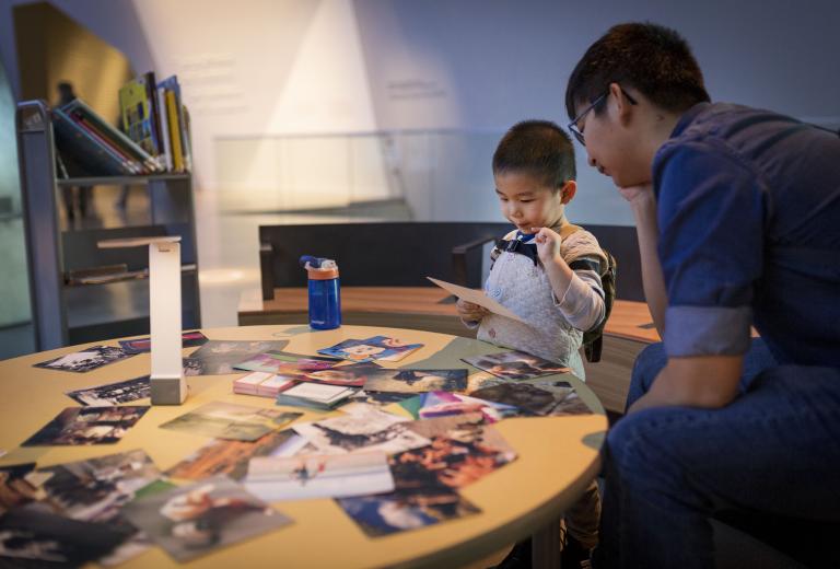 Un enfant interagit avec des photos étalées sur une table. Un adulte est assis à côté de lui. Visibilité masquée.