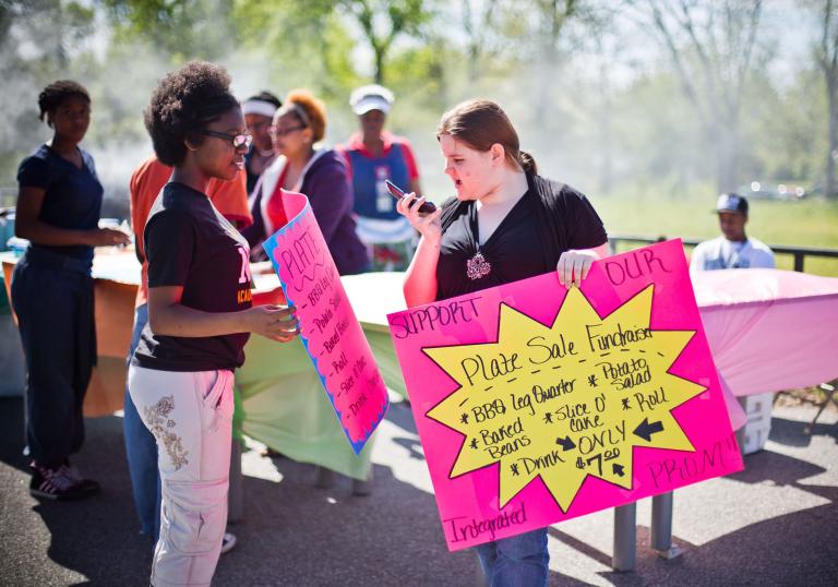Deux femmes tiennent des affiches qui annoncent une collecte de fonds pour la vente d’assiettes. La femme à droite parle dans un téléphone qu’elle tient devant son visage, plutôt qu’à son oreille.
