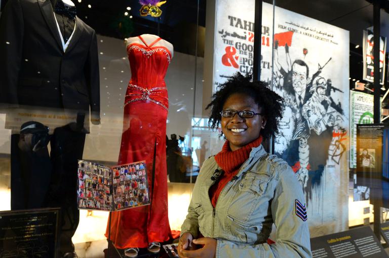 Une femme souriante se tient devant une robe de bal rouge et un smoking noir exposés sur des mannequins. La robe et le costume sont derrière une vitrine. Visibilité masquée.