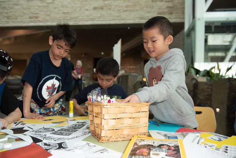 Un enfant plonge dans un panier de matériel de bricolage pendant que d'autres travaillent à leurs projets à une table. Visibilité masquée.