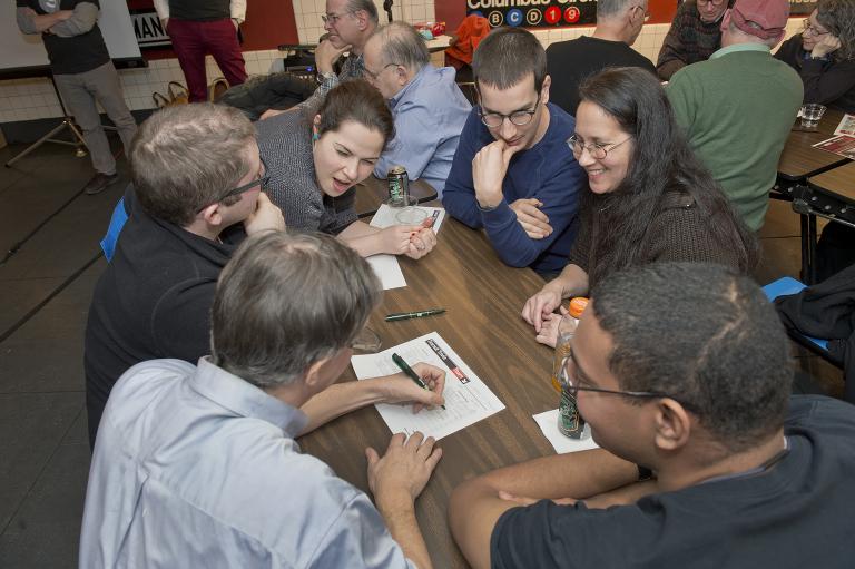 Des gens enthousiasmés regroupés autour d’une table. Un homme écrit sur une feuille de papier. Visibilité masquée.