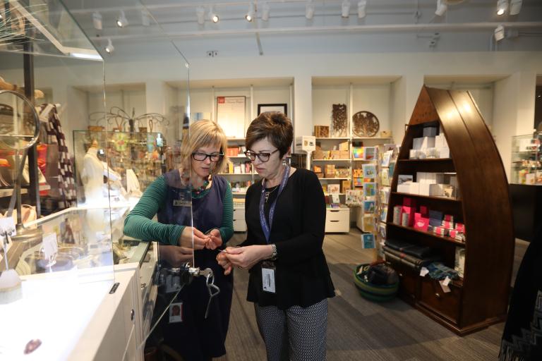 Deux femmes regardent un bijou. Elles sont debout dans un magasin rempli de marchandises colorées. Visibilité masquée.