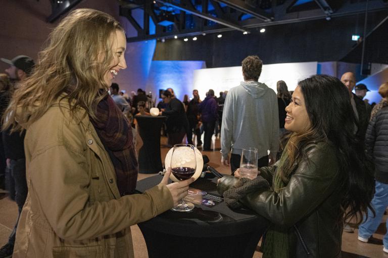 Deux femmes souriantes boivent du vin dans une grande salle remplie de gens. Visibilité masquée.