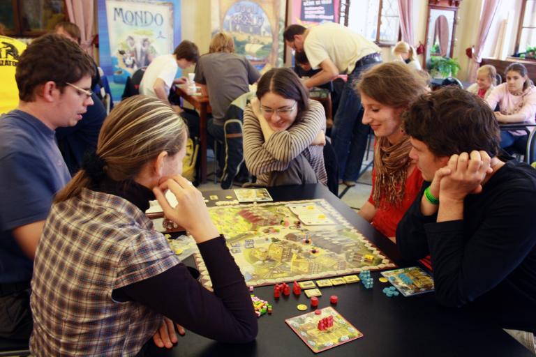 Trois jeunes femmes et deux jeunes hommes sont assis autour d’une table et jouent un jeu de société. En arrière-plan, d’autres personnes jouent à des jeux à d’autres tables. Visibilité masquée.