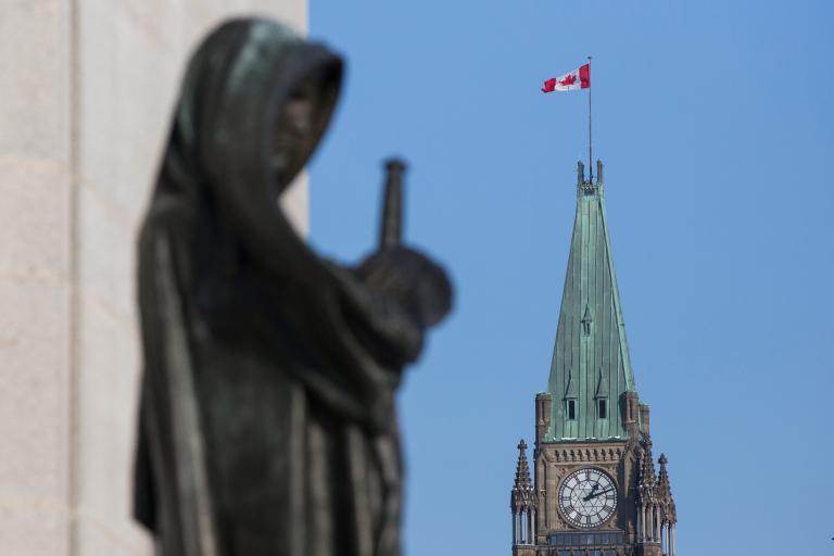 Un drapeau canadien flotte au sommet d’une grande tour. Au premier plan se dresse la statue d’une femme voilée portant une épée. Visibilité masquée.