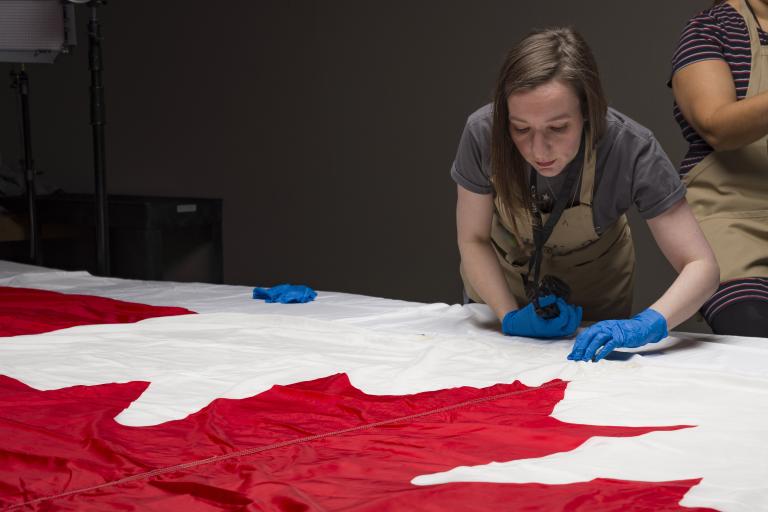 Une femme portant des gants de protection et un tablier inspecte un grand drapeau du Canada, posé à plat sur une table.