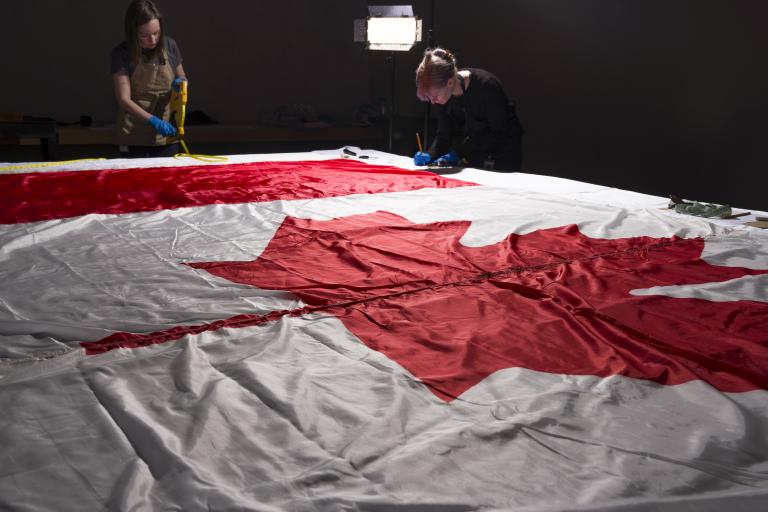 Deux femmes portant des gants et des tabliers de protection autour d’un grand drapeau du Canada, posé à plat sur une table.