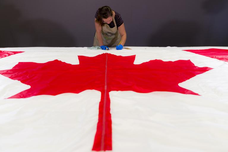 Une femme portant des gants de protection et un tablier prend des notes derrière un drapeau du Canada, posé à plat sur une table à l’avant-plan.