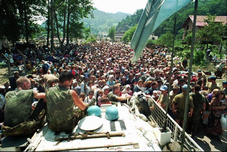 Deux soldats en uniforme sont assis sur un grand véhicule, surplombant une foule dense de personnes qui s’étend au loin. Visibilité masquée.