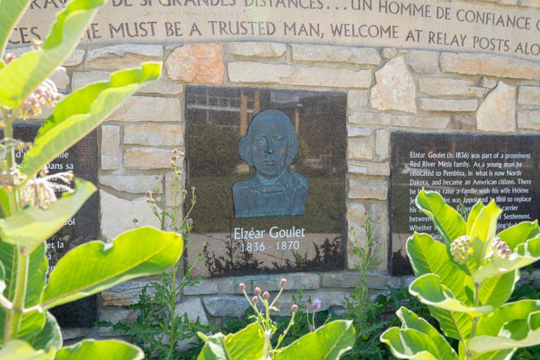 Un mur de pierre avec une écriture décorative sculptée, des panneaux d’information et un portrait en bas-relief d’un homme au-dessus des mots « Elzéar Goulet 1836-1870 ».