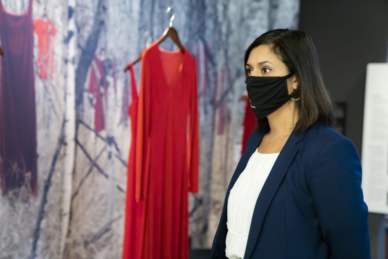 Une femme portant un masque noir se tient devant une exposition du Musée composée de robes rouges suspendues devant un fond de forêt. Visibilité masquée.