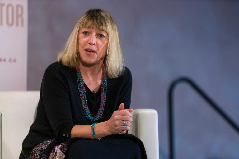 Une femme assise sur un fauteuil, les mains jointes, regarde vers l’avant d’un air pensif, comme si elle répond à une question. Visibilité masquée.