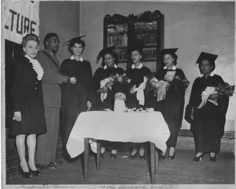 Une image en noir et blanc de Viola Desmond debout à côté d’un homme en uniforme. Debout à leur gauche, cinq femmes portent des coiffes et des robes de finissante et tiennent des fleurs. Une femme accepte un parchemin roulé de l’homme en uniforme.