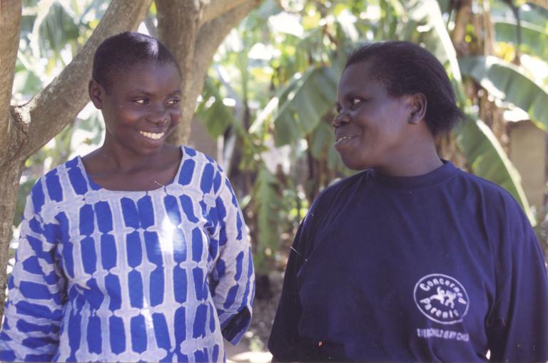 Deux femmes, debout devant des arbres tropicaux, se regardent et sourient. La femme de droite porte un chandail qui porte le logo de la Concerned Parents Association.