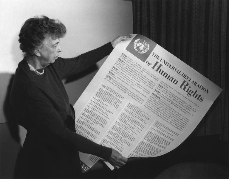 Une personne tient une grande feuille de papier recouverte de texte et dont le gros titre est « The Universal Declaration of Human Rights. » Visibilité masquée.