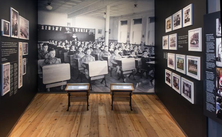 Une exposition muséale qui comprend de petits pupitres d’école devant un grand fond photographique d’enfants autochtones assis à leur pupitre dans une classe. Visibilité masquée.