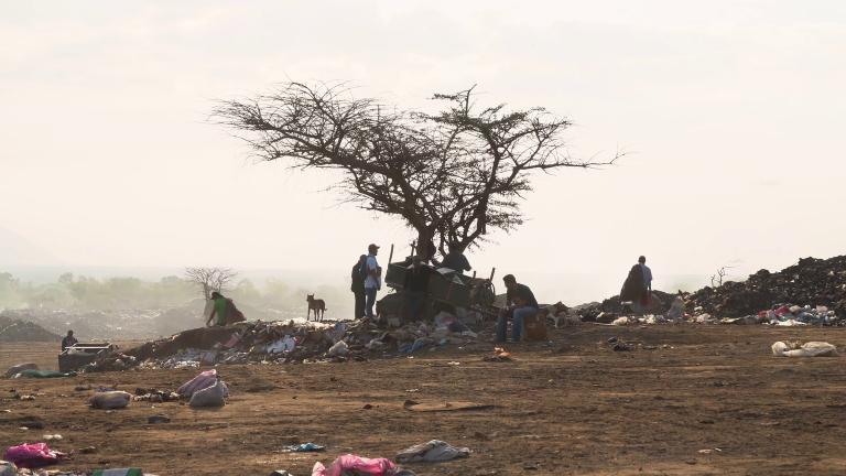 Plusieurs personnes et un chien assis et debout dans une décharge. Un arbre est au centre de l'image et des déchets sont éparpillés sur le sol. Visibilité masquée.
