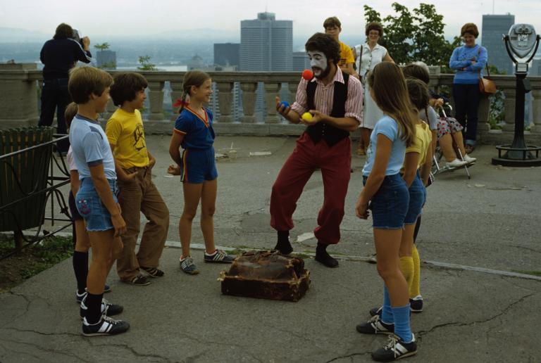 Un homme au visage peint jongle en plein air, sous le regard d'enfants souriants. Une silhouette de ville apparaît au loin derrière lui.