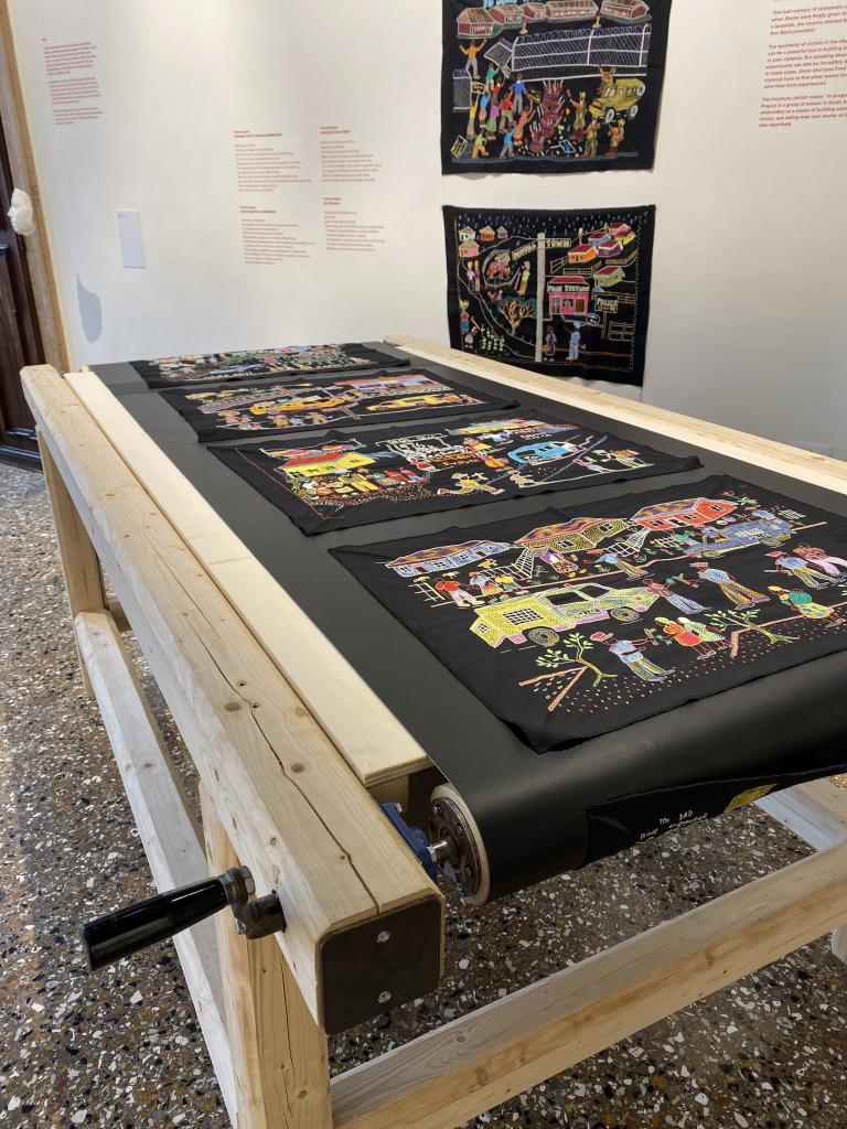 Une série de panneaux de broderie colorés sur du tissu noir, exposés sur une table de convoyeur en bois à manivelle et sur le mur d’un musée.