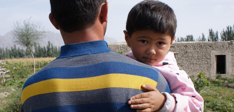 Un jeune enfant porté par un adulte regarde en arrière par-dessus l’épaule de l’adulte. Visibilité masquée.
