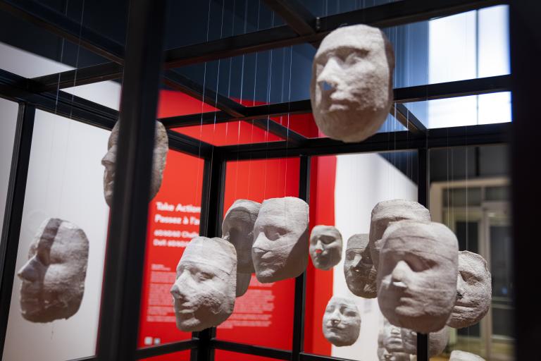 Des moulages blancs de visages de femmes sont suspendus par des fils aux barreaux d’acier d’une grande cage. Visibilité masquée.