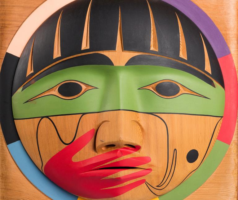 Une boîte en bois, sur laquelle est sculpté un visage dont la bouche est recouverte d’une main peinte en rouge. Visibilité masquée.