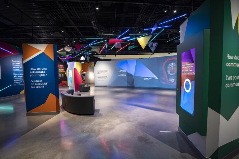 Une galerie de musée où on voit des oeuvres d'art projetées sur de grands écrans et formes géométriques colorées. Visibilité masquée.