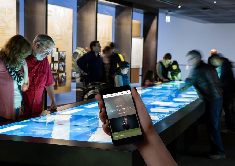 Une main tient un téléphone portable. Sur le téléphone se trouvent les mots « Niveau 4 » et « Briser le silence », et une personne parlant en langage des signes. En arrière-plan, des personnes regardent une grande table rectangulaire avec un écran tactile. Visibilité masquée.
