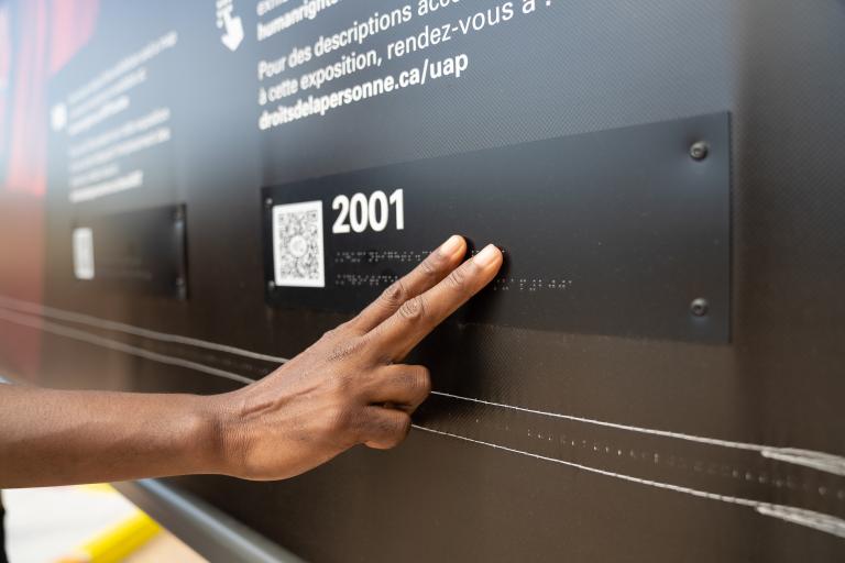 Une main touche des lettres en braille dans une exposition muséale. Visibilité masquée.