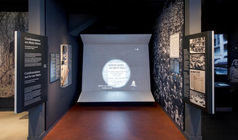 Une exposition muséale comprenant des photos, du texte et un grand écran vidéo avec des sous-titres et une interprétation en langue des signes.