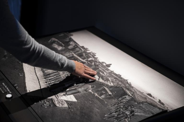 Une main touche une grande image en relief par endroits, dont les personnes et bâtiments représentés ont une texture visible.