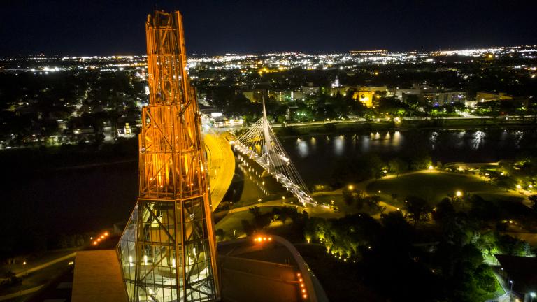 Une tour illuminée en orange vue d’en haut avec en arrière-plan un paysage urbain de nuit où on voit aussi une rivière et un pont.