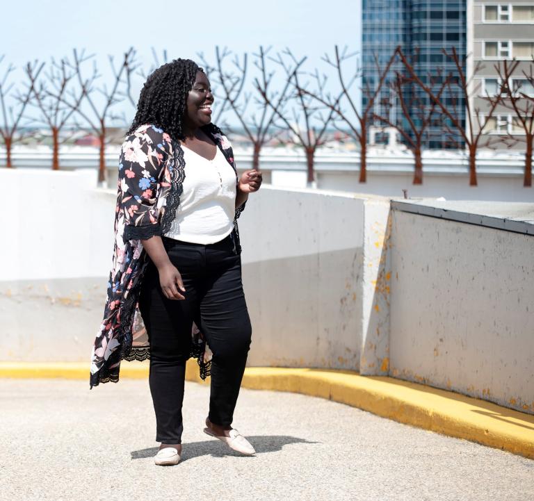 Une femme noire souriante est debout devant une rangée d'arbres. Elle regarde vers sa gauche. Visibilité masquée.