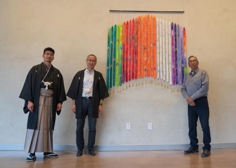 Trois hommes debout près d’une œuvre composée de mille grues multicolores en papier plié. Deux des hommes portent des vêtement traditionnels japonais. L’autre homme porte une chemise couleur denim et une cravate bolo.