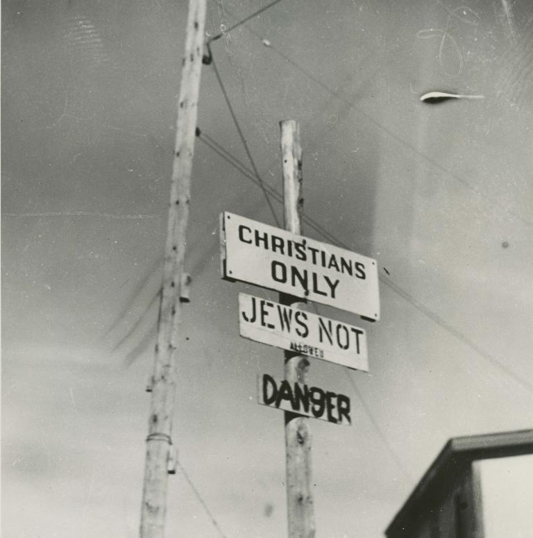 Photo noir et blanc de trois écriteaux fixés sur un poteau, où l’on peut lire en anglais « Chrétiens seulement », « Interdit aux Juifs » et « Danger ». Visibilité masquée.