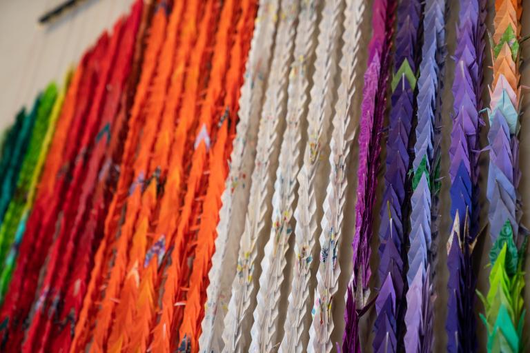 Grues multicolores en origami pressées les unes contre les autres verticalement et suspendues au mur. Visibilité masquée.