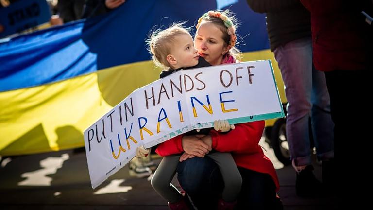 Un enfant tient un panneau de protestation qui dit, en anglais : « Poutine, ne touche pas à l’Ukraine. » L’enfant est assis sur les genoux de la personne qui s'occupe de lui. Derrière eux se trouve un drapeau ukrainien. Visibilité masquée.