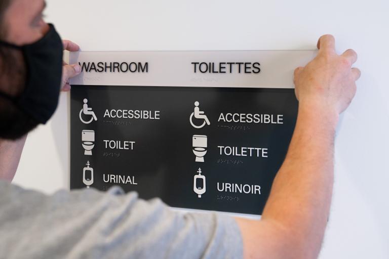A man holds up a sign that says "Washroom" as it is mounted to a wall. Underneath the title, the words "accessible," "toilet" and "urinal" are accompanied by descriptive icons. Visibilité masquée.