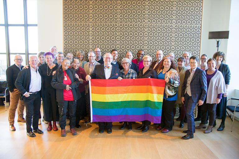  Vingt-huit membres du conseil d’ administration du Fonds Purge LGBT et du Comité consultatif du Monument national LGBTQ2+ posant ensemble et tenant un drapeau arc-en-ciel, lors de la première session de visualisation du Monument en 2019. Visibilité masquée.
